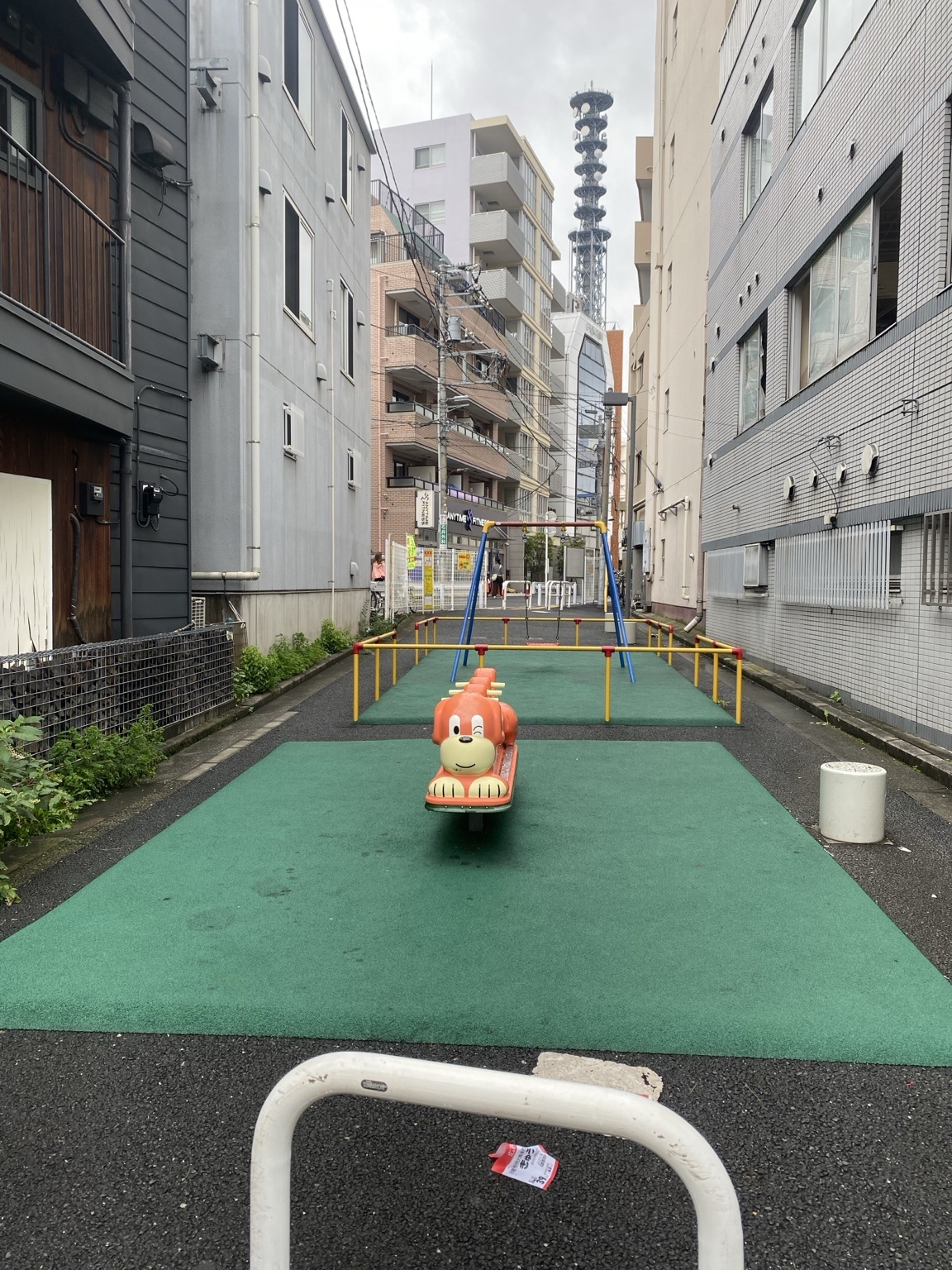 ビルに挟まれた細長い公園。緑のマットに犬のスプリング遊具とブランコ、奥にエニタイムフィットネスの看板と鉄塔が見える